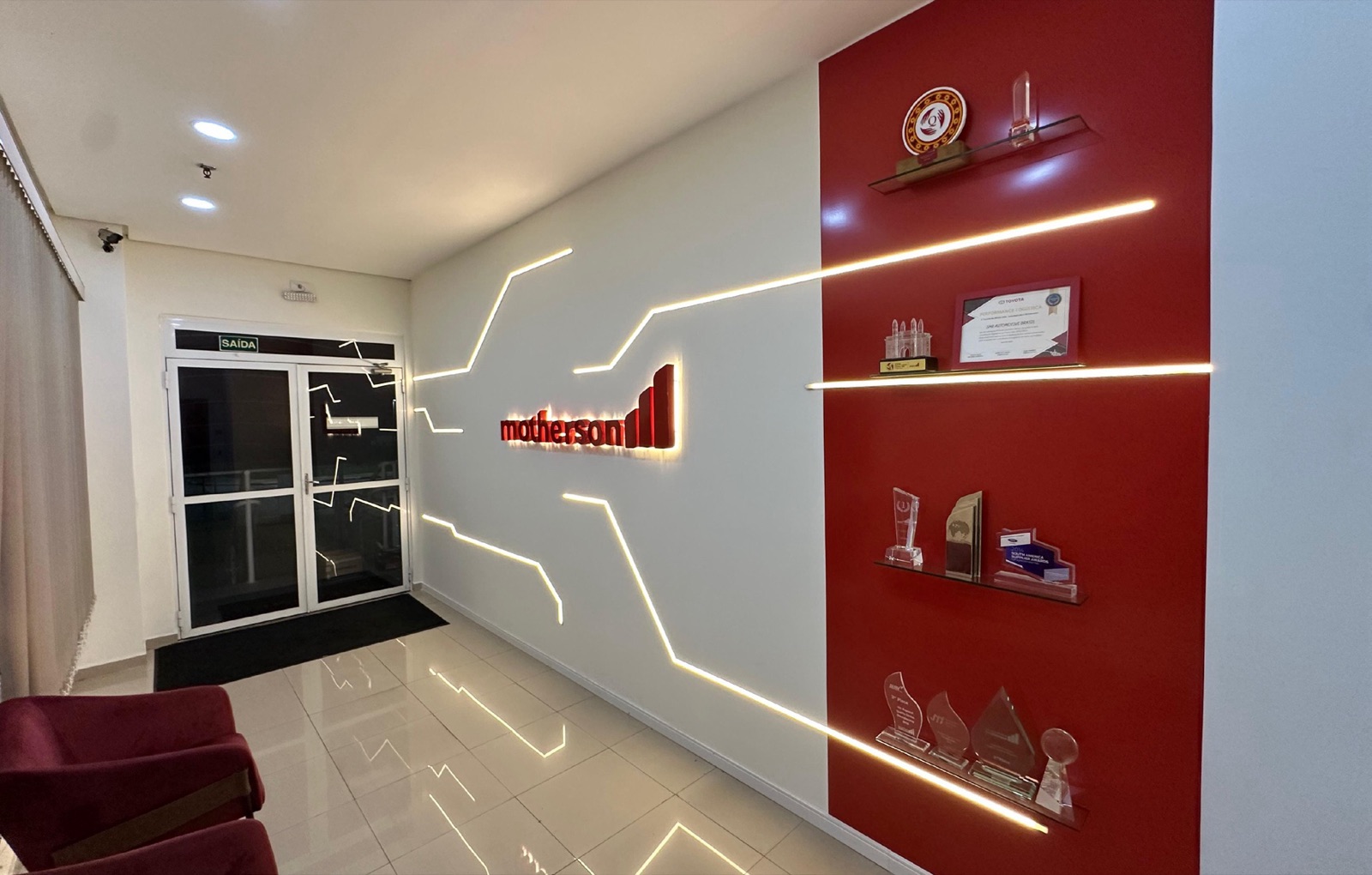 Recepção executada da SMR Automotive, com parede vermelha, iluminação linear e prateleiras de premiações